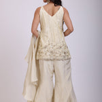 A woman modeling a cream-colored traditional outfit with matching wide-leg trousers, featuring a sleeveless embroidered top and a draped dupatta, set against a plain light gray background.