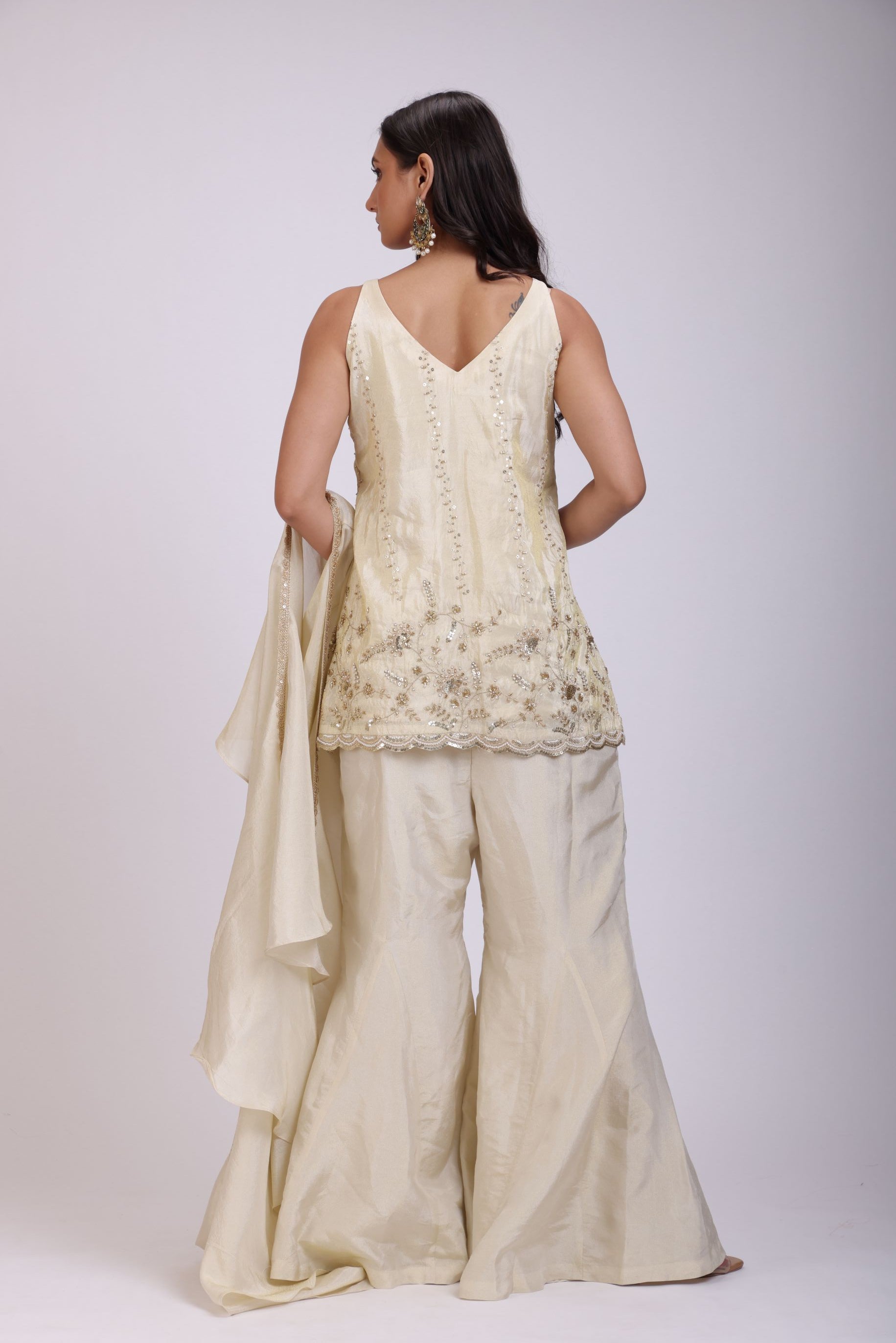 A woman modeling a cream-colored traditional outfit with matching wide-leg trousers, featuring a sleeveless embroidered top and a draped dupatta, set against a plain light gray background.
