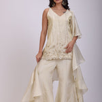 A woman modeling a cream-colored traditional outfit consisting of a heavily embroidered kurta and matching wide-leg trousers, posed against a plain light gray studio background.