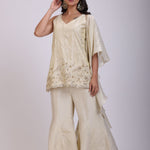 A woman modeling a cream-colored traditional outfit consisting of a sleeveless embroidered top and matching wide-leg pants, set against a plain light gray studio background.